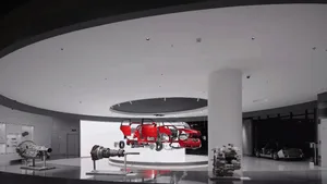 Modern exhibition hall with a red car chassis and body suspended in the center, surrounded by automotive components on stands under a curved white ceiling.