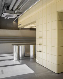 Modern commercial interior with a pale yellow perforated metal wall, integrated floating shelves, cylindrical stainless steel bench, exposed concrete ceiling with visible ductwork and air conditioning unit.