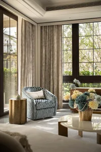 A contemporary living room corner featuring a blue and white checkered upholstered armchair, a textured gold cylindrical side table, a large marble coffee table with a floral arrangement in a brass vase, and floor-to-ceiling windows with neutral curtains.