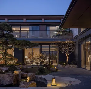 Modern Japanese-style courtyard at dusk featuring a stone-lined pond, pine tree, bare trees, stone path, and glowing lanterns against a contemporary house with large glass windows.