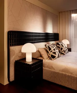 Modern bedroom interior with a large black grid-patterned headboard, textured beige fabric upholstery, mushroom table lamps, and black and beige geometric pillows.