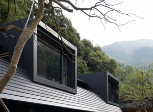 Modern black metal house with large windows nestled among green mountains and trees, viewed from below with foreground tree branches.