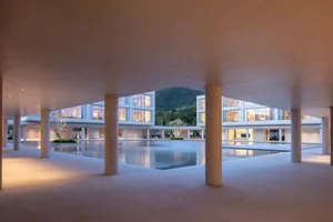 View from a concrete colonnade overlooking a large rectangular reflecting pool surrounded by modern white buildings with floor-to-ceiling glass and cylindrical concrete pillars, with a green mountain in the background.