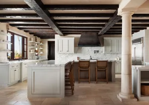 Bright and airy kitchen interior with a large white marble island, three woven leather bar stools, white shaker cabinets, marble backsplash, dark reclaimed wood ceiling beams, and a stone column.