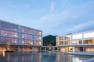 Modern two-story glass and concrete villa with illuminated interiors surrounding a large reflecting pool courtyard at twilight, with mountains in the background.
