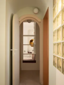View down a pastel-colored hallway featuring a curved arched doorway, white spherical ceiling lights, and a luminous wall of yellow glass blocks.