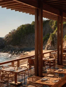 Outdoor dining area with teak tables and woven rattan chairs on a wooden deck overlooking a sandy beach, rocky cliffs, and the ocean under a clear sky.