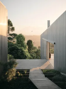 View from inside a modern concrete house looking out onto a wooden deck terrace at golden hour with forested hills in the background.