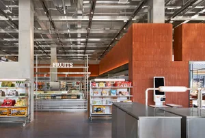 Modern supermarket interior showing stainless steel checkout counters, orange-brown corten steel wall panels, metal shelving units, and an exposed industrial ceiling with visible ductwork and lighting.