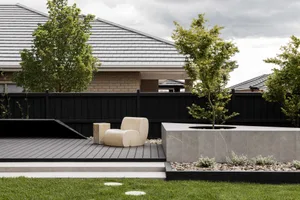 Modern backyard featuring a sculptural cream lounge chair on a dark timber deck, a large concrete planter with a young tree, black fence, and contemporary house with tiled roof.
