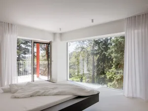 Minimalist bedroom with low platform bed dressed in white linen, sheer white curtains, and large windows overlooking a lush green forest.