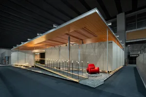 現代展覽亭擁有木質天棚、白色紋理牆面、黑色金屬欄杆，以及一張鮮豔紅色單椅置於礫石平台上，搭配戲劇性照明。