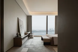Modern luxury bedroom interior featuring a large harbor view window, walnut media console, textured area rug, and woven bench at the foot of the bed.
