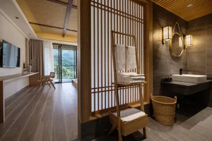 Modern Japanese-style bathroom featuring a wooden slat room divider, vessel sink on stone countertop, woven towel ladder, and large glass doors opening to a green mountain view.