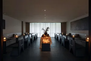 Interior view of a minimalist fine dining restaurant featuring dark stone floors and walls, built-in banquette seating, angular dark wood tables and chairs, and numerous lit candles creating warm ambient lighting, with large floor-to-ceiling windows at the far end.