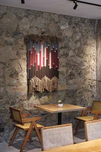 Dining area featuring a large colorful woven wall tapestry with fringe against a textured stone wall, accompanied by wooden cane-back chairs and a light wood table.
