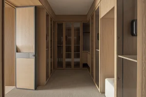 Modern walk-in closet featuring light oak cabinetry, glass-fronted storage, built-in seating, and neutral carpet flooring.