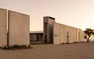 Modern concrete house named Eyrie with tall chimney, stacked firewood, and long horizontal walls at sunset in a rural setting.