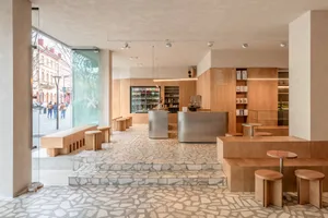 Modern minimalist café interior with oak wood counters, stainless steel service bar, and irregular stone tile flooring, large glass windows overlooking the street.