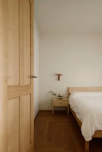 Minimalist bedroom interior featuring light oak wood flooring, matching bed frame and nightstand, white bedding, and a modern terracotta wall sconce with a small pine branch arrangement.