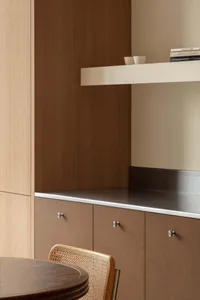 Close-up of a modern dining area featuring warm walnut lower cabinets with round silver knobs, a stainless steel countertop, light oak upper cabinetry, and a cane-backed dining chair in the foreground.