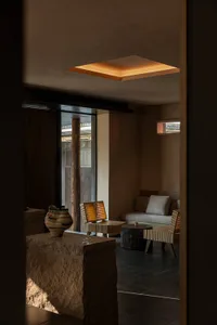 Modern minimalist lounge with raw stone blocks, woven rattan chairs, dark tiled floor, and a glowing square skylight in an earthy-toned interior.