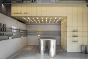 Modern minimalist beauty boutique interior featuring a central stainless steel cylindrical display island, pale yellow tiled walls, concrete flooring, and product shelves under a grid ceiling with linear lighting.