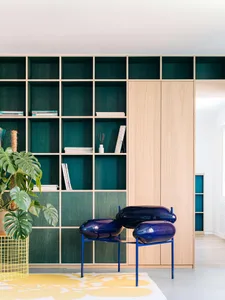 Modern living room with teal velvet built-in shelving, light wood cabinetry, a glossy blue sculptural chair, a large monstera plant, and a yellow floral rug.