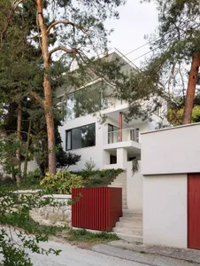 Modern white house with large glass windows, white balconies, concrete stairs, and vibrant red metal accents on a wooded hillside.