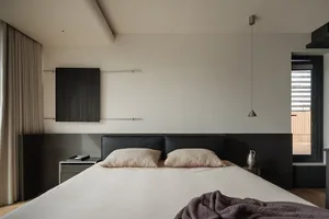 Modern minimalist bedroom with a large bed dressed in white linen and beige textured pillows, set against a dark wood floating headboard panel. A single pendant light hangs above, with a black wood wall cabinet mounted on the left wall.