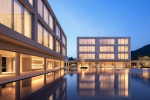 Modern minimalist building complex with large glass windows and concrete structure surrounding a large rectangular reflection pool at twilight, with mountains in the background.