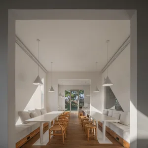 Long, bright minimalist dining room with two parallel white tables, built-in oak banquettes with white cushions, wishbone chairs, and pendant lights, viewed through a rectangular opening.