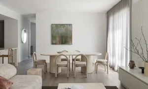 Modern minimalist dining room featuring a large travertine oval table, six curved upholstered chairs, abstract green painting, and sheer curtains allowing natural light.
