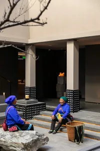 Two elderly Miao women wearing vibrant blue traditional costumes and headwraps sit on stone steps in a modern concrete and stone courtyard at night, illuminated by warm lighting.