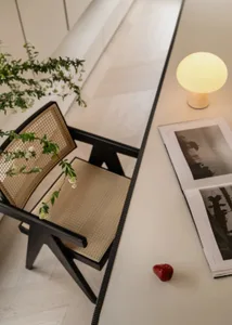 High-angle view of a black wooden dining chair with natural rattan woven seat and backrest, placed beside a long white dining table featuring a glowing spherical table lamp, an open photography book, and a small red pepper, with green branches in the foreground.