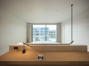 Modern minimalist bedroom interior with light oak desk, black articulated lamp, freestanding white bathtub, and large glass doors opening to a mountain and lake view.