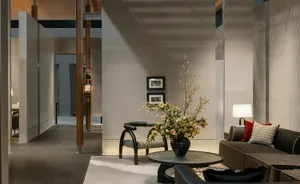Modern minimalist living room featuring exposed concrete walls, a dark L-shaped sofa, a round glass coffee table, a sculptural black chair, and a large dramatic floral arrangement in a black vase.