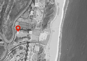 Black and white aerial photograph of a coastal resort complex with a red location pin marking one of the buildings, bordered by a sandy beach and the sea.