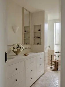 Modern minimalist bathroom with white limestone vanity, integrated sink, dark metal hardware, recessed wall niches, and a wooden chair visible in the adjacent space.