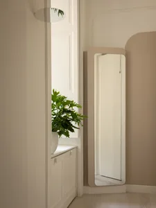 Minimalist entryway corner featuring a tall full-length mirror with a matte beige frame, a white arched wall detail, and a large potted monstera plant on a white cabinet by the window.