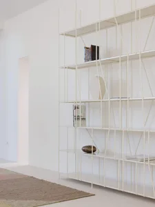 Modern white metal grid bookshelf against a white wall, displaying books, ceramic vases, and sculptural objects, with a textured area rug in the foreground.