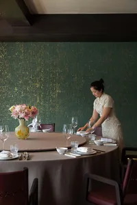Elegant Chinese fine dining room featuring a large round table set with porcelain and crystal, set against a deep emerald green textured wall with gold leaf accents, with a server in a cream lace qipao arranging tableware.
