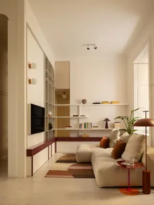 Modern minimalist living room with off-white walls, built-in white shelving, terracotta-red accents, a beige modular sofa with brown and beige cushions, a striped area rug, and a large potted plant.