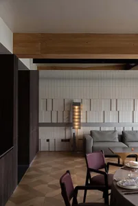 Modern dining room featuring burgundy leather chairs, a round dining table set with tableware, a grey sofa, herringbone wood flooring, and a feature wall with beige textured tiles and a wooden beam overhead.
