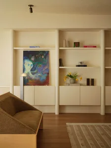 Modern minimalist living room with light oak flooring, built-in white shelving, a brown ribbed fabric armchair, colorful abstract painting, and decorative objects.