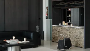 Modern dark living room featuring a black leather sofa, burl wood cabinet with white marble top, black rocking ottoman, concrete pedestal with cat sculpture, and dramatic dark wood paneling.
