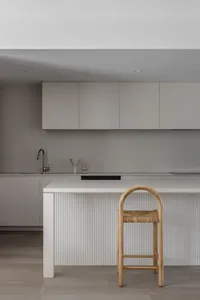 Modern minimalist kitchen with grey flat-panel cabinets, white fluted kitchen island, wooden rattan bar stool, and matte brass faucet.