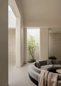 Modern minimalist living room featuring a large curved grey sectional sofa, travertine stone walls and floor, and an indoor courtyard with a Japanese maple tree.