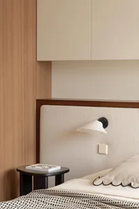 Modern minimalist bedroom corner showing a cream corduroy upholstered headboard, dark wood trim, light wood wall paneling, black wall sconce, and round black side table with a book.