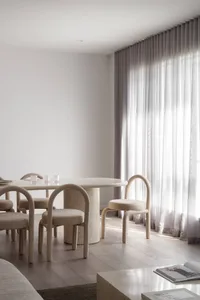 Minimalist dining room featuring a round travertine table, curved beige upholstered chairs, light wood flooring, and floor-to-ceiling sheer curtains with bright natural light.